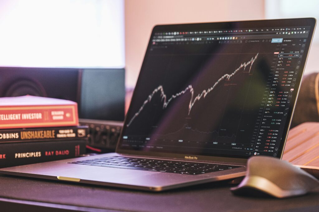 Laptop showing stock market charts with investing books including The Intelligent Investor and Principles by Ray Dalio